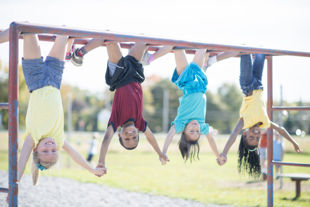 A multi-ethnic group of elementary age children are hanging upside down on the monkey bars at the school playground. They are holding hands and smiling at the camera.